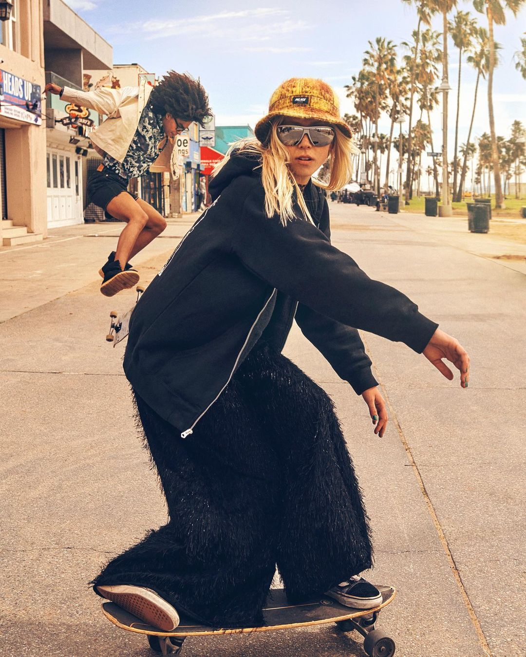 Blonde woman in black hoodie and fringed pants skateboarding on an outdoor path with another skater in the background.