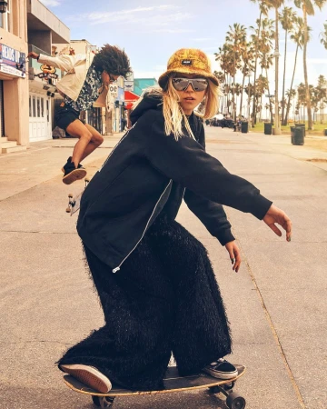 Blonde woman in black hoodie and fringed pants skateboarding on an outdoor path with another skater in the background.
