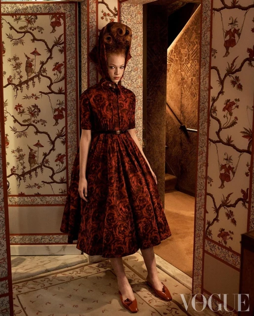 Fashion editorial shot of a red-haired model wearing a vintage-inspired red print dress and matching shoes, posed in a patterned room.