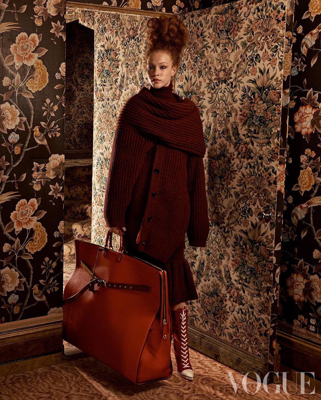 A young woman with red hair wears a burgundy sweater and dress, holding a large leather bag, posing indoors against floral wallpaper.