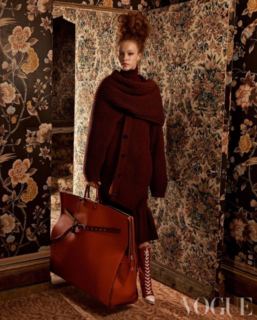 A young woman with red hair wears a burgundy sweater and dress, holding a large leather bag, posing indoors against floral wallpaper.