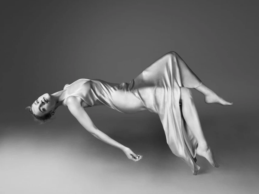 Elegant woman in flowing dress floats mid-air against a smooth grey backdrop in a monochrome studio-style fashion portrait.