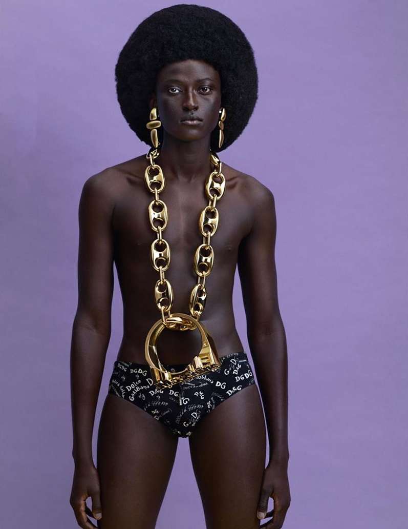 Model with large afro hairstyle wearing oversized gold chain necklace and printed briefs against purple background