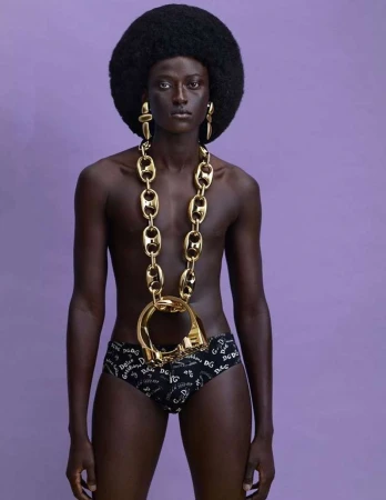 Model with large afro hairstyle wearing oversized gold chain necklace and printed briefs against purple background