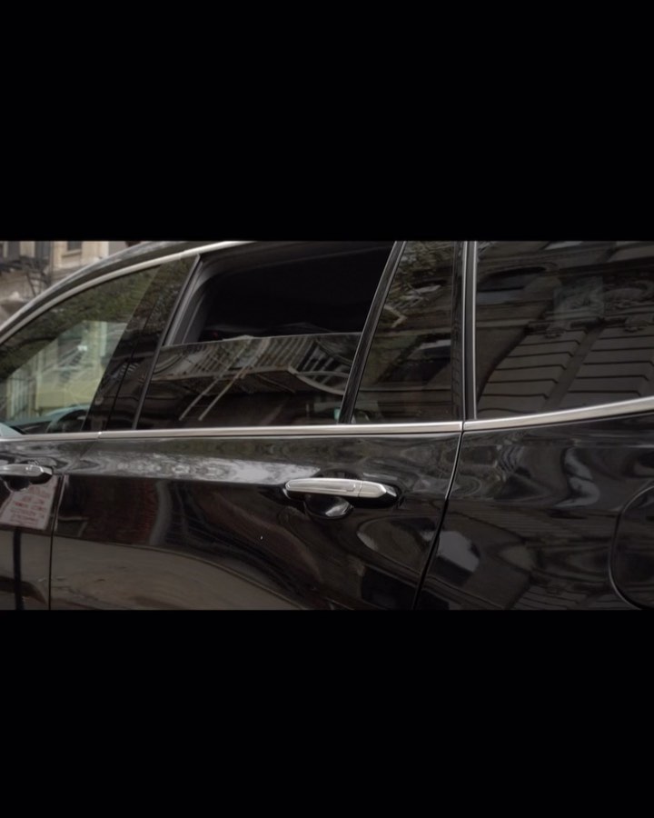 Side view of a shiny black SUV parked outdoors, reflecting the surroundings in its windows. The vehicle is parked on a street.