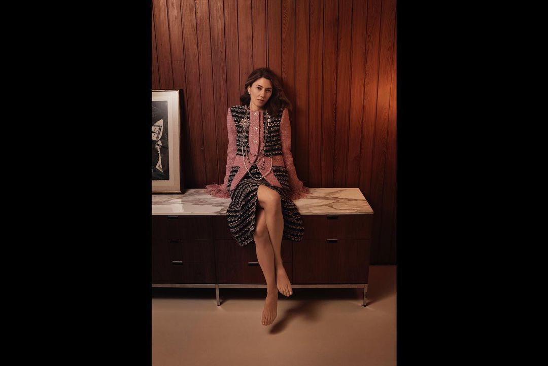 A woman in a patterned dress and vest sits on a cabinet in a well-lit interior with wood paneling and a framed picture on the cabinet.