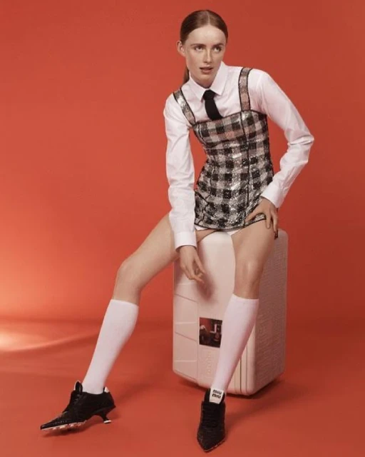 A slender adult female is seated indoors on a white cube, wearing a plaid minidress, long-sleeved shirt, tie, knee socks, and black heeled shoes.