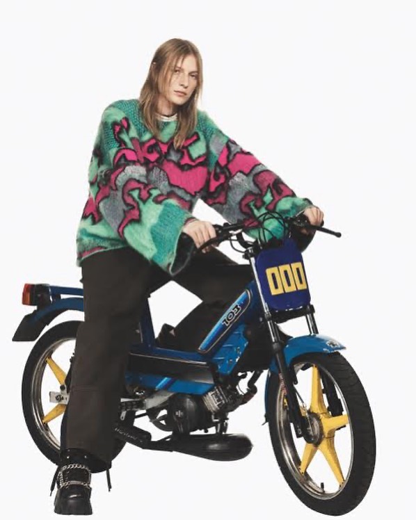 Woman in a colorful sweater rides a blue moped.  A studio shot features bright lighting and a white backdrop.