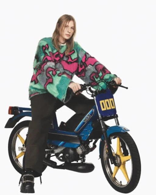 Woman in a colorful sweater rides a blue moped.  A studio shot features bright lighting and a white backdrop.