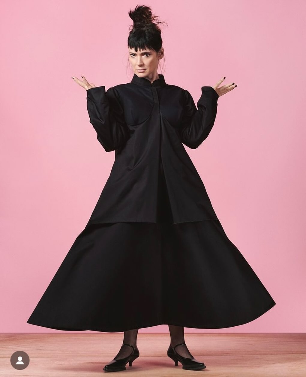 Elegant adult woman in black dress posing with hands up against a pink background inside a photo studio, low heels, stockings.