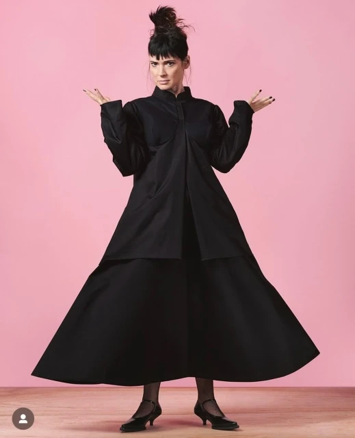 Elegant adult woman in black dress posing with hands up against a pink background inside a photo studio, low heels, stockings.