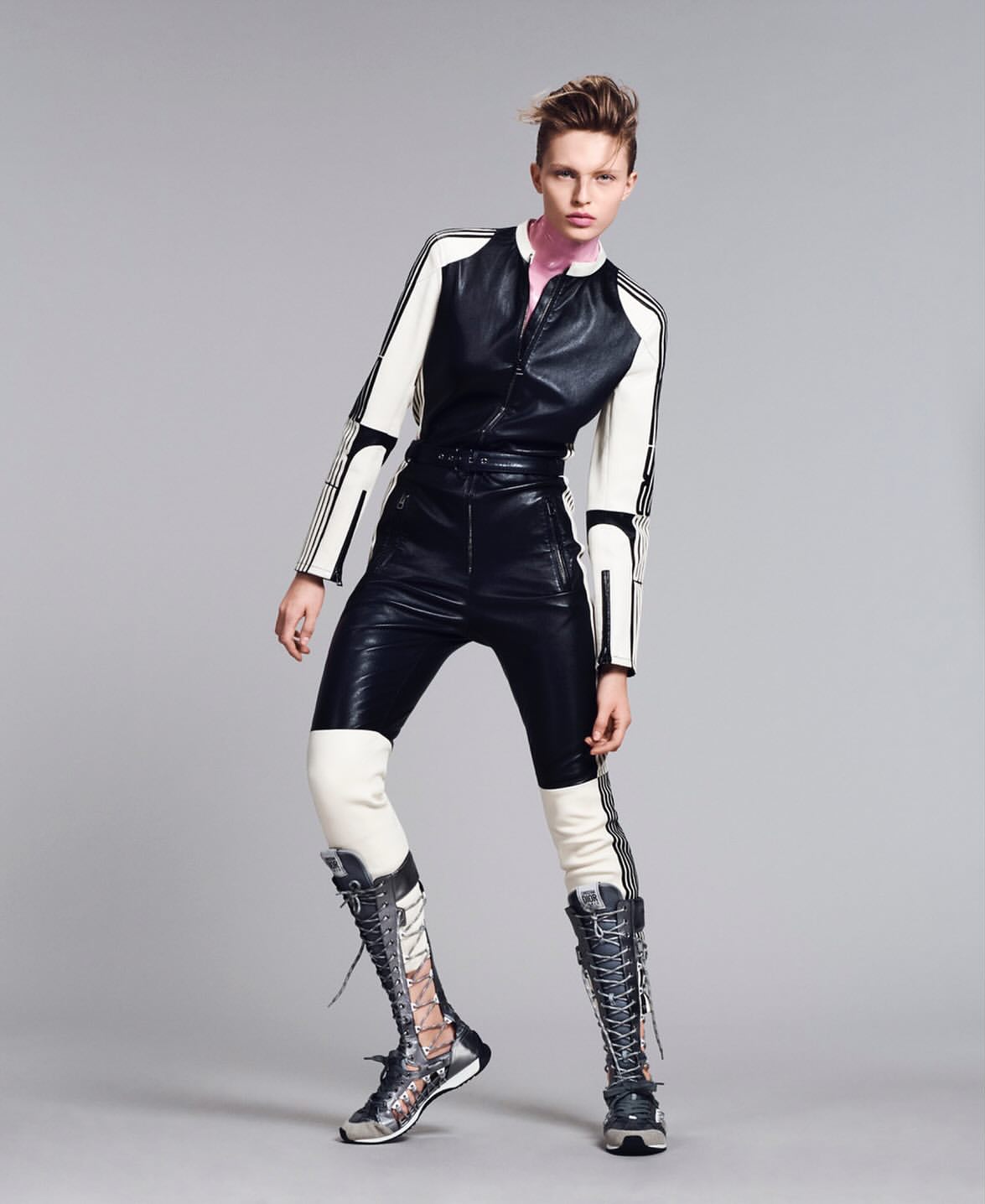 Fashion editorial shot of a stylish woman posing in black and white leather jumpsuit with lace up boots against a grey studio backdrop.