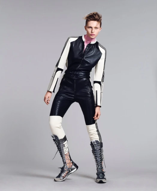 Fashion editorial shot of a stylish woman posing in black and white leather jumpsuit with lace up boots against a grey studio backdrop.