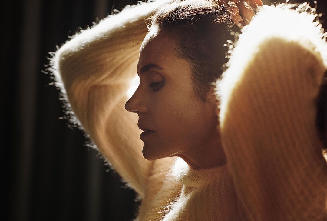 Profile of a woman with hands in her hair wearing a soft sweater in a softly lit indoor setting, fashion and beauty photography.