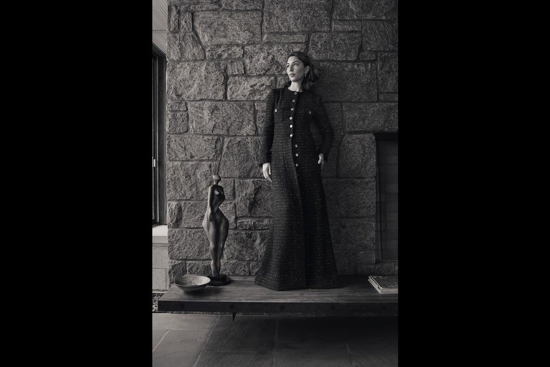 Monochrome portrait of a woman in a long tweed coat posing against a stone wall with a sculpture and neutral-toned interior.
