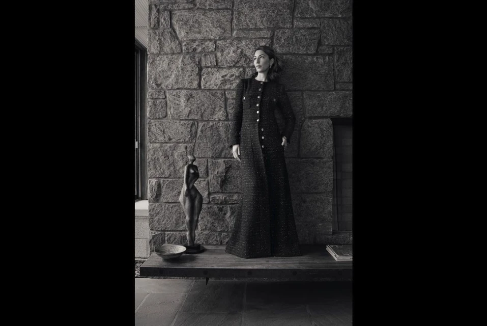 Monochrome portrait of a woman in a long tweed coat posing against a stone wall with a sculpture and neutral-toned interior.