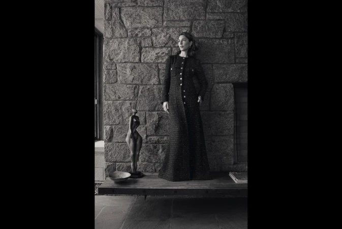Monochrome portrait of a woman in a long tweed coat posing against a stone wall with a sculpture and neutral-toned interior.