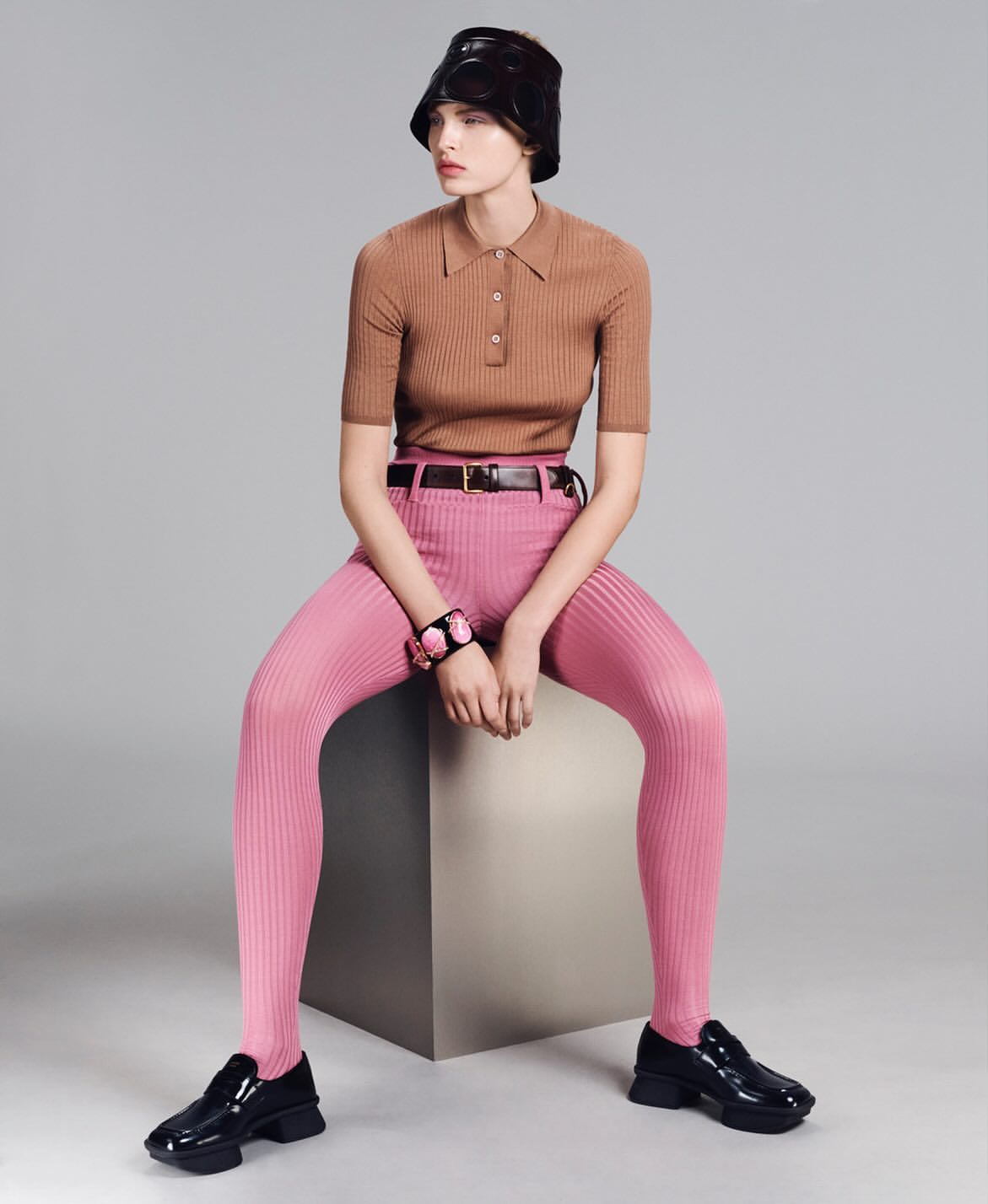 A female fashion model poses seated on a cube, wearing a brown polo shirt, pink leggings, black shoes, and a black hat against a grey backdrop.