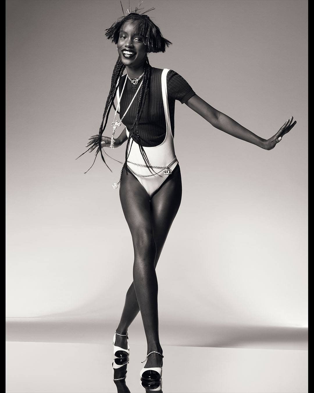 Monochrome studio shot of a slender African woman in a white bodysuit and black top, posing with outstretched arms and smiling.