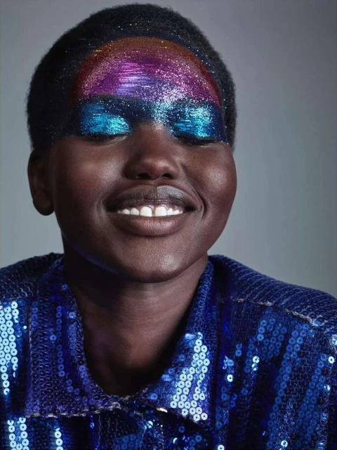 Close-up portrait of a dark-skinned woman with glitter makeup on her eyelids and forehead, wearing a blue sequined jacket, smiling softly.