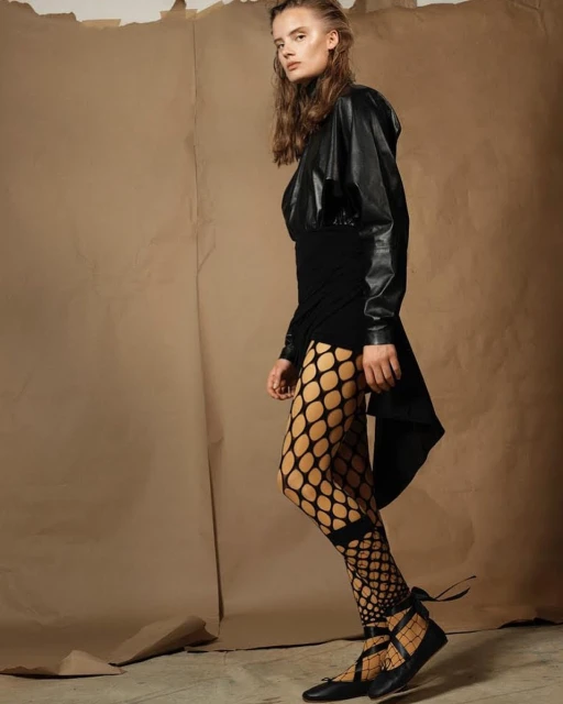 A young woman in a black dress and fishnet tights poses in a studio with a backdrop of brown craft paper and wears black ballet flats.