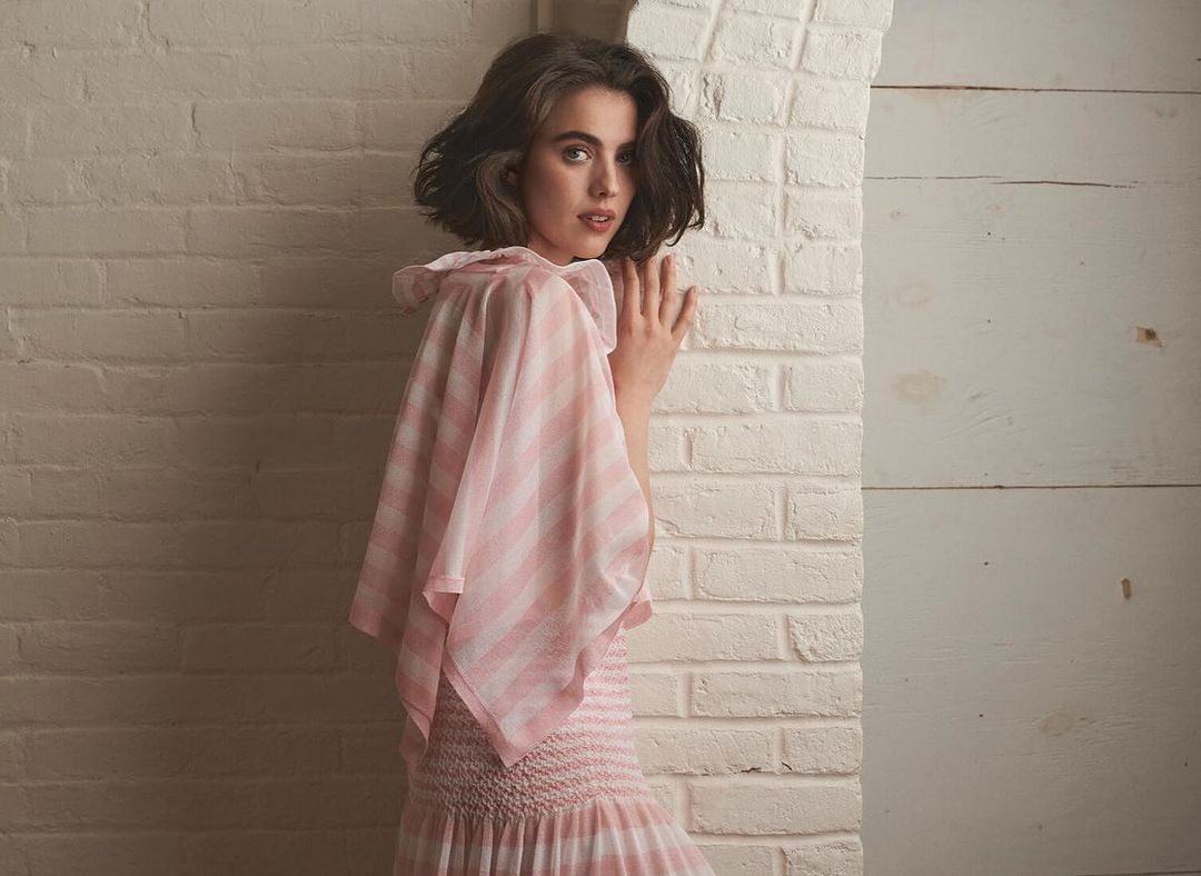 Young woman with shoulder-length brunette hair poses next to a white brick wall in a pink and white striped dress with frilled neckline.