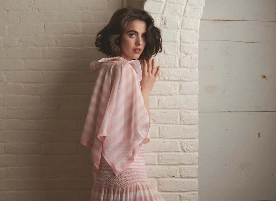 Young woman with shoulder-length brunette hair poses next to a white brick wall in a pink and white striped dress with frilled neckline.