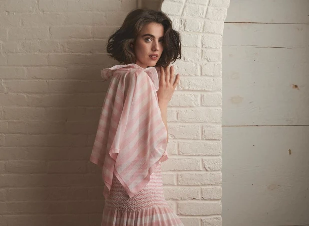 Young woman with shoulder-length brunette hair poses next to a white brick wall in a pink and white striped dress with frilled neckline.