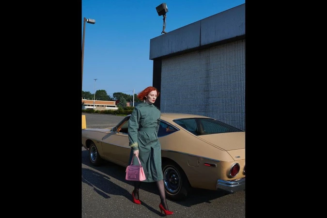 Red-haired woman walking in a green dress and red heels past a tan car outdoors