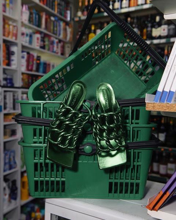 Emerald green Bottega Veneta Madame chain mules resting in stacked green plastic shopping baskets in a store aisle setting.