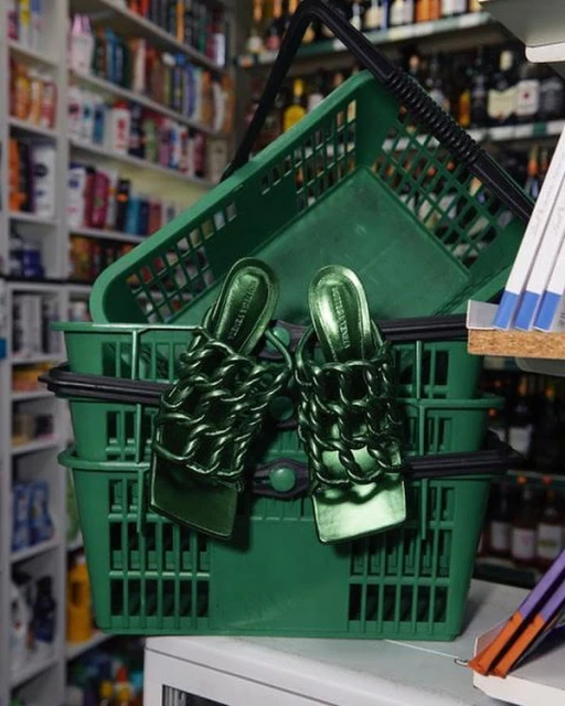 Emerald green Bottega Veneta Madame chain mules resting in stacked green plastic shopping baskets in a store aisle setting.