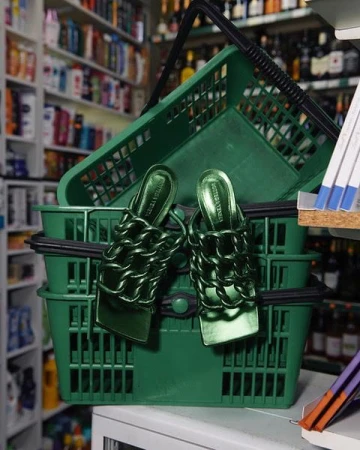 Emerald green Bottega Veneta Madame chain mules resting in stacked green plastic shopping baskets in a store aisle setting.