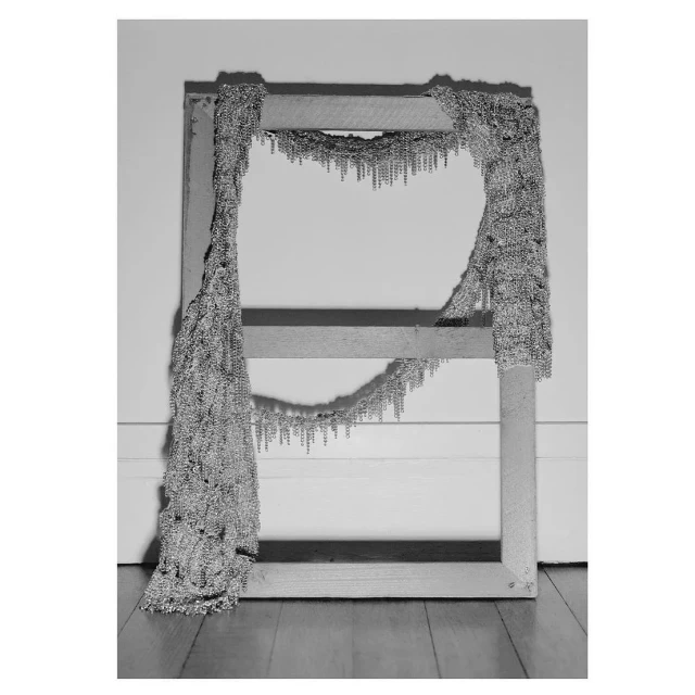 Monochrome image of a wooden frame draped with metallic strands, casting shadows on a wall with a wooden floor, studio lighting.