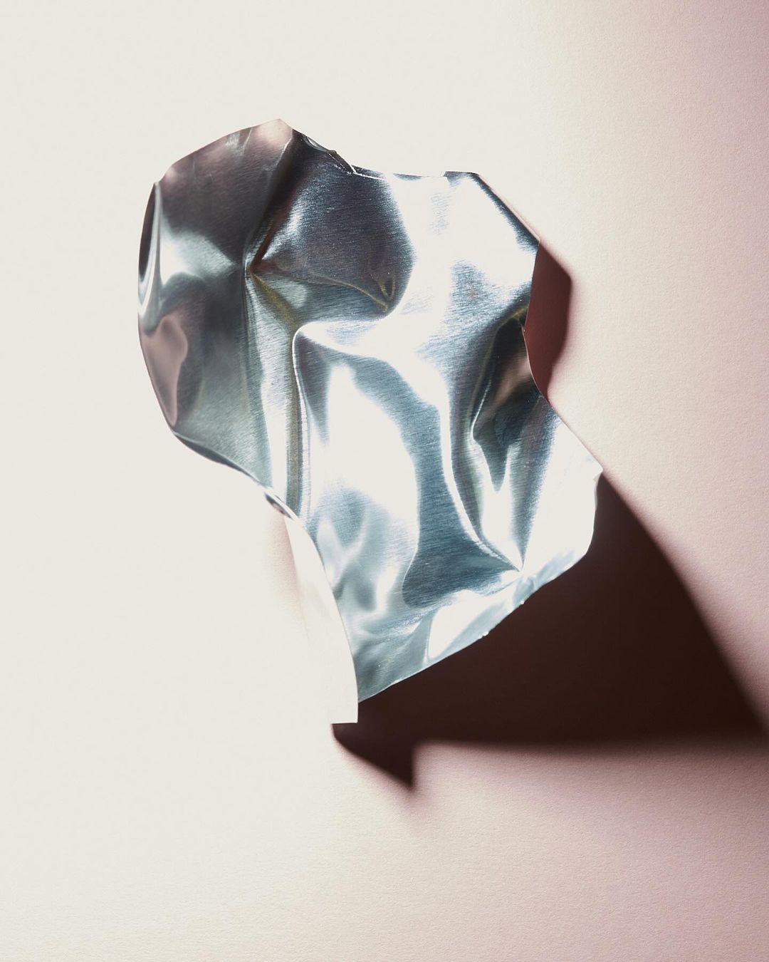 Close-up of a crumpled, iridescent metallic sheet against a soft pink backdrop, casting a dark shadow, creating a modern abstract.