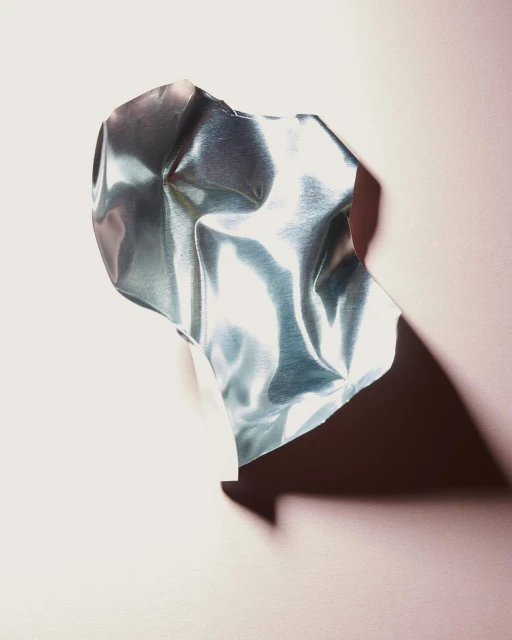 Close-up of a crumpled, iridescent metallic sheet against a soft pink backdrop, casting a dark shadow, creating a modern abstract.