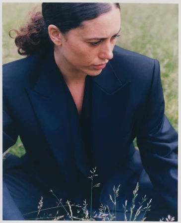 Woman in a dark tailored blazer sitting outdoors in a grassy field with wild grass stems in the foreground