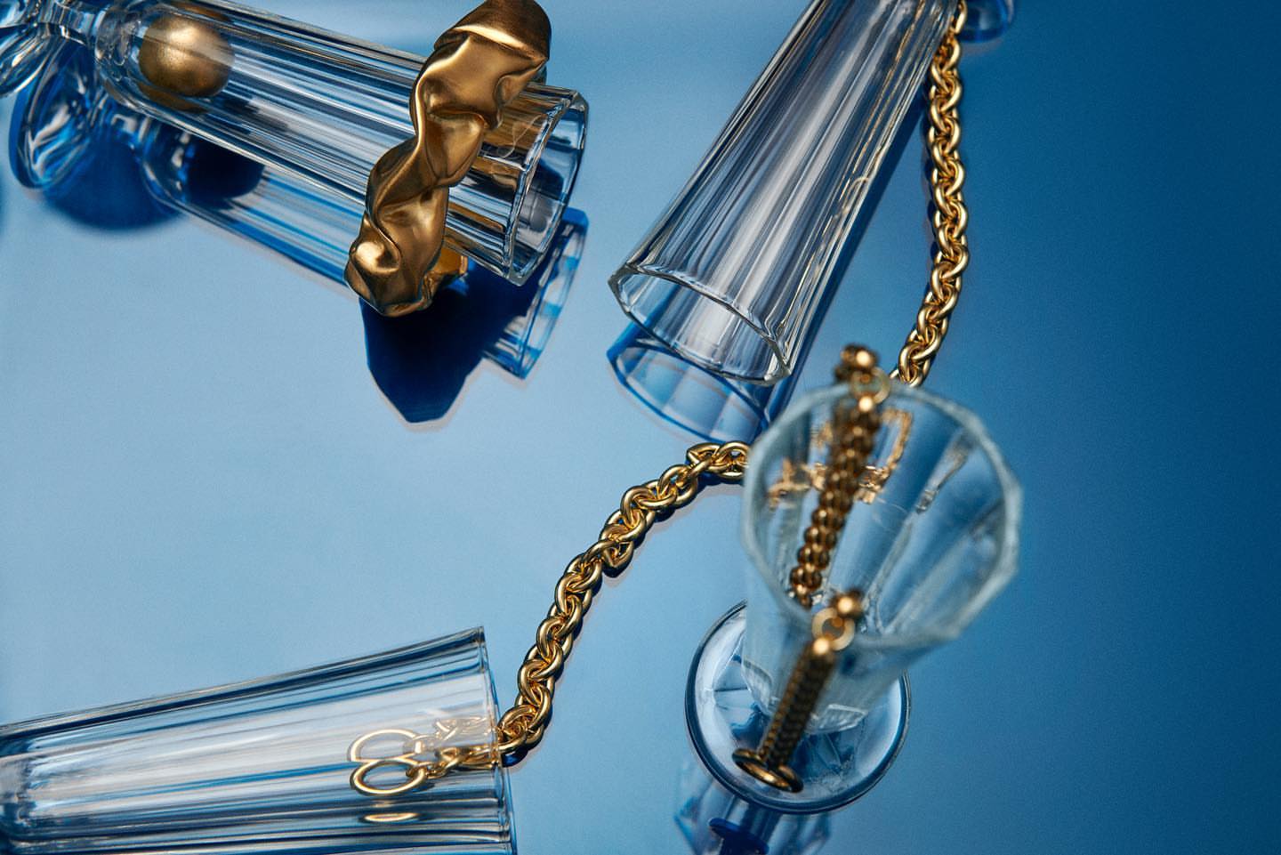 Still life photo of clear ribbed drinking glasses connected by a gold chain, set against a graduated light to dark blue background.