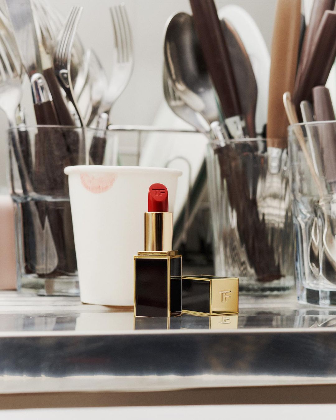 Still life of Tom Ford lipstick tube with open cap on silver tray near white paper cup and cutlery in glass jars.