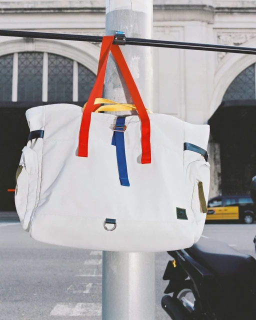 White tote bag with red, yellow, and blue straps hanging from a metal pole on a city street with a building and vehicle in the background.