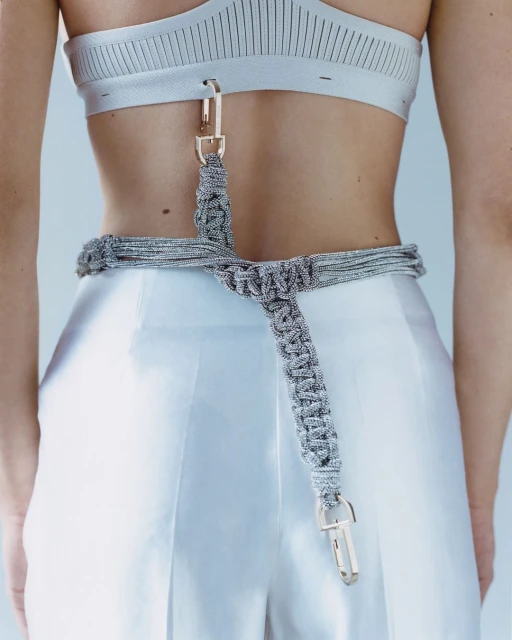 Close-up on a woman with a silver sports bra and white pants, accessorized with a silver macrame belt and gold metal hardware.