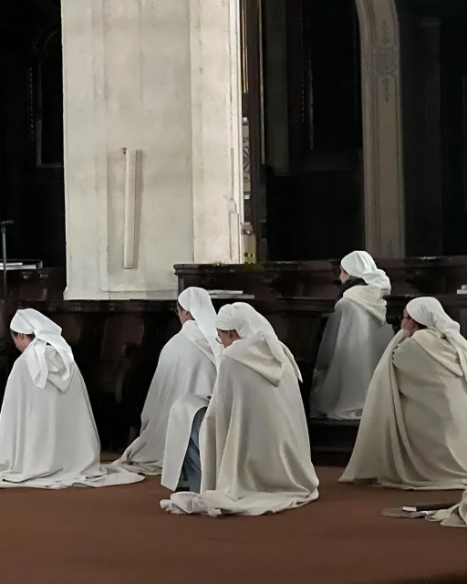 Group of people kneeling in white robes inside church, heads covered by white veils, creates a quiet and contemplative scene.