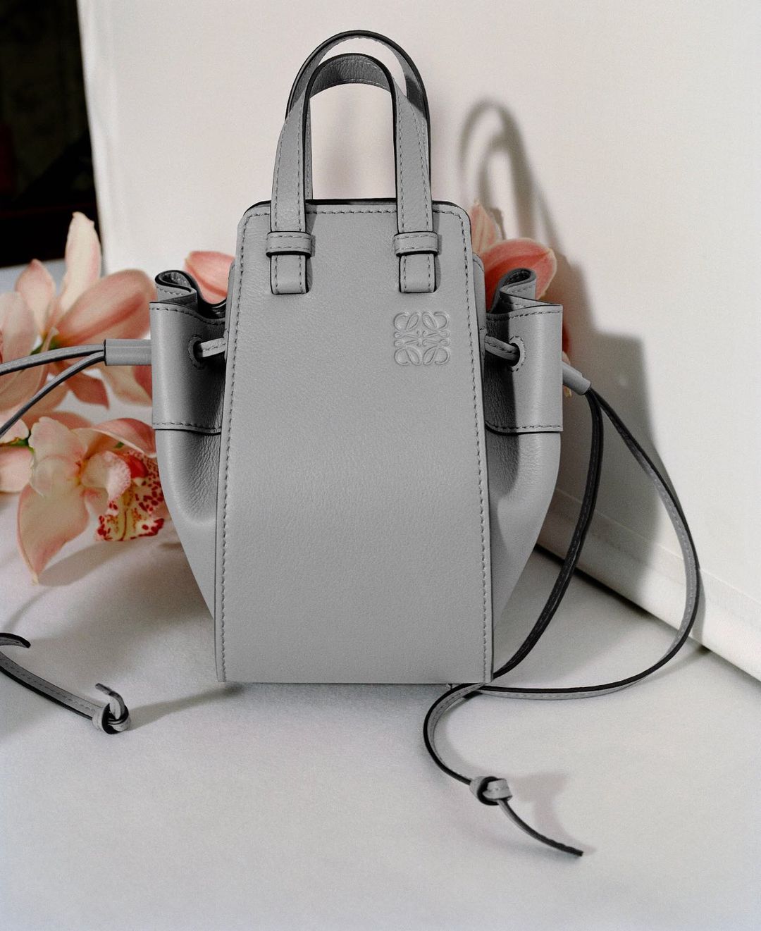 Grey leather handbag with thin straps and a debossed logo set against a white backdrop with pink floral accents