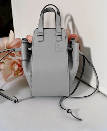 Grey leather handbag with thin straps and a debossed logo set against a white backdrop with pink floral accents