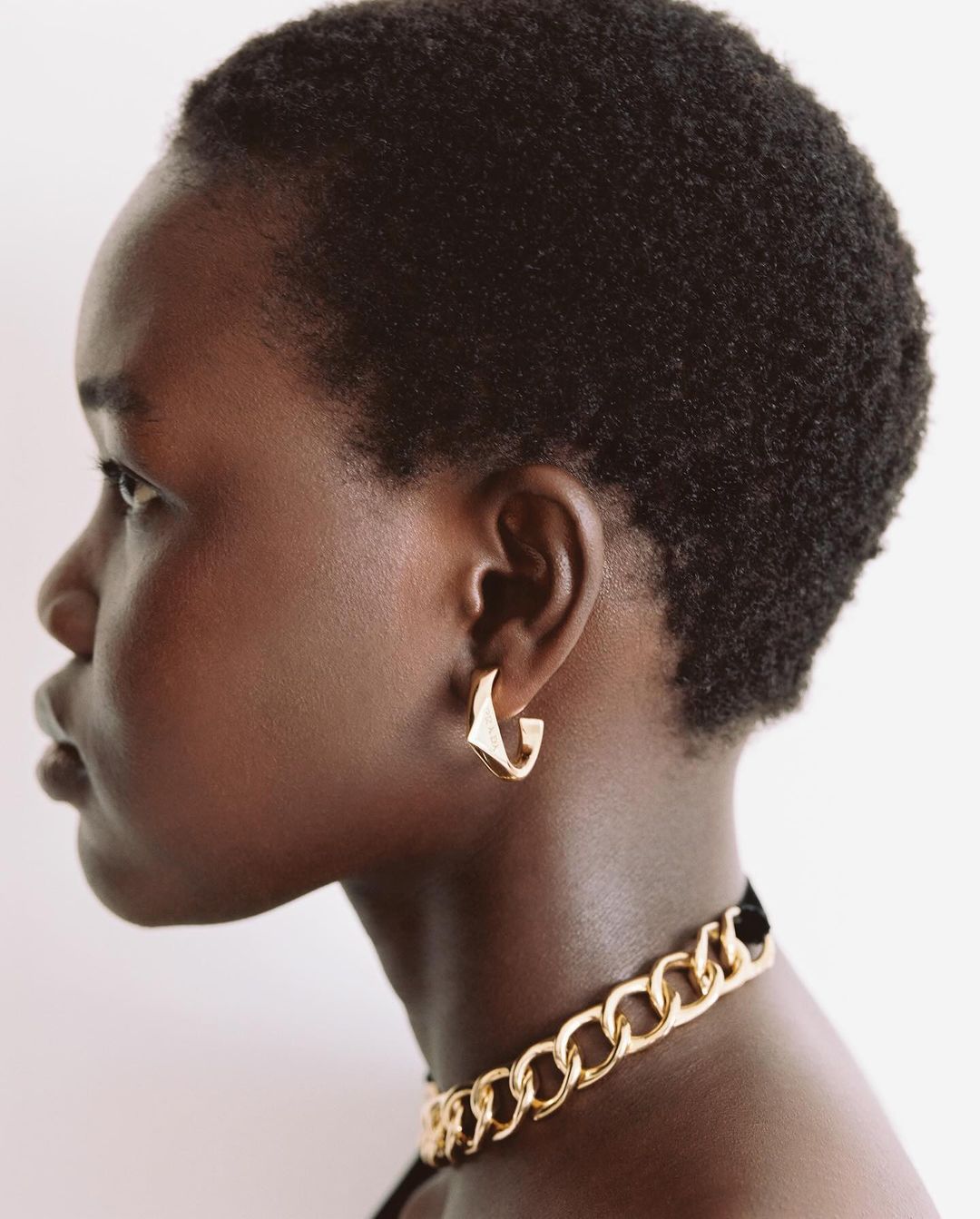 Profile view of a Black woman with short natural hair wearing gold chain necklace and earrings against a neutral backdrop.