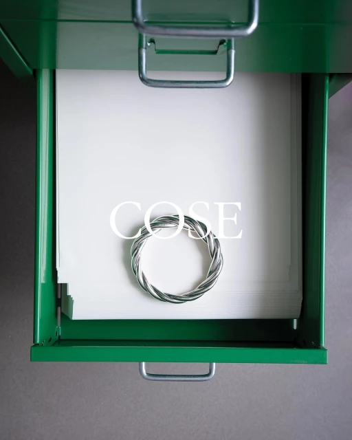 Close-up of a green metal drawer opened to reveal stacked white paper sheets and a circular wire design in an indoor setting.
