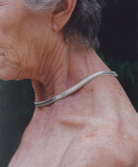 Older adult with short gray hair wearing a silver necklace in an outdoor setting with a blurred natural green background. 