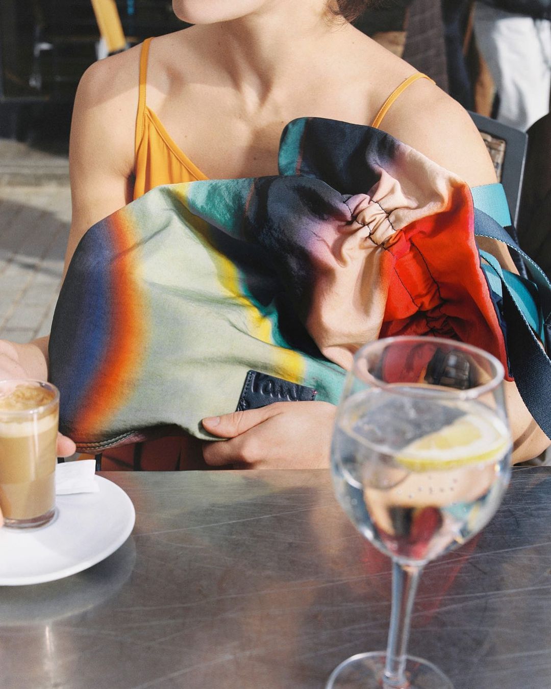 Woman sits at an outdoor cafe table with a colorful abstract print purse, water glass with lemon, and small espresso.