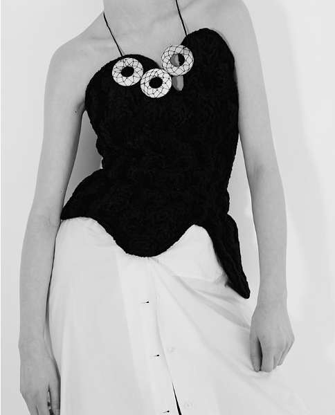 Model wearing a textured black strapless top with circular white necklace accessories over a white skirt against a plain background