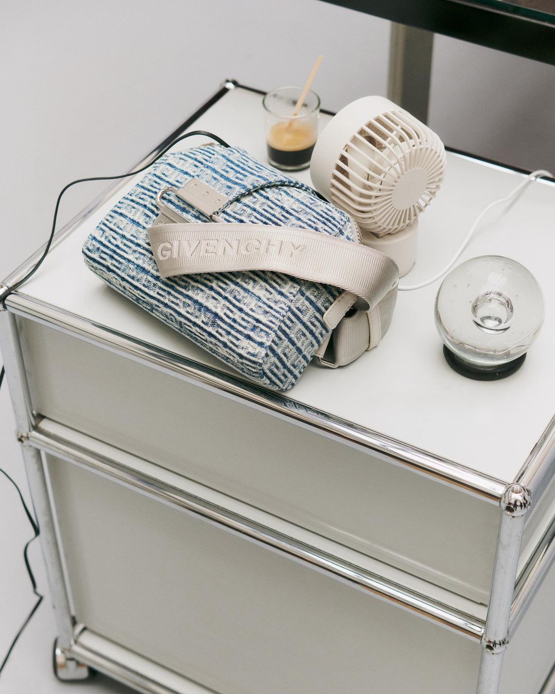 Givenchy handbag on a white table next to a small electric fan, glass, and metal side table in a bright, indoor setting.