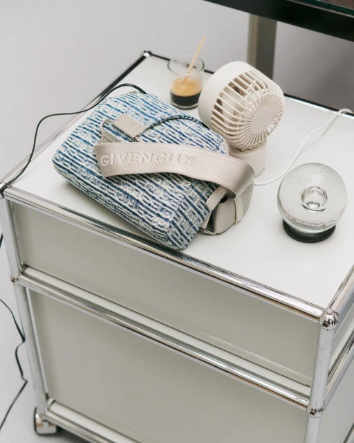 Givenchy handbag on a white table next to a small electric fan, glass, and metal side table in a bright, indoor setting.